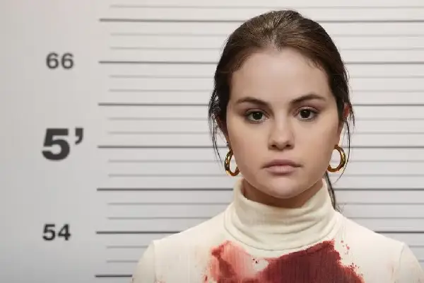 Mabel standing solemnly in front of a police lineup wall, her white shirt stained with blood.